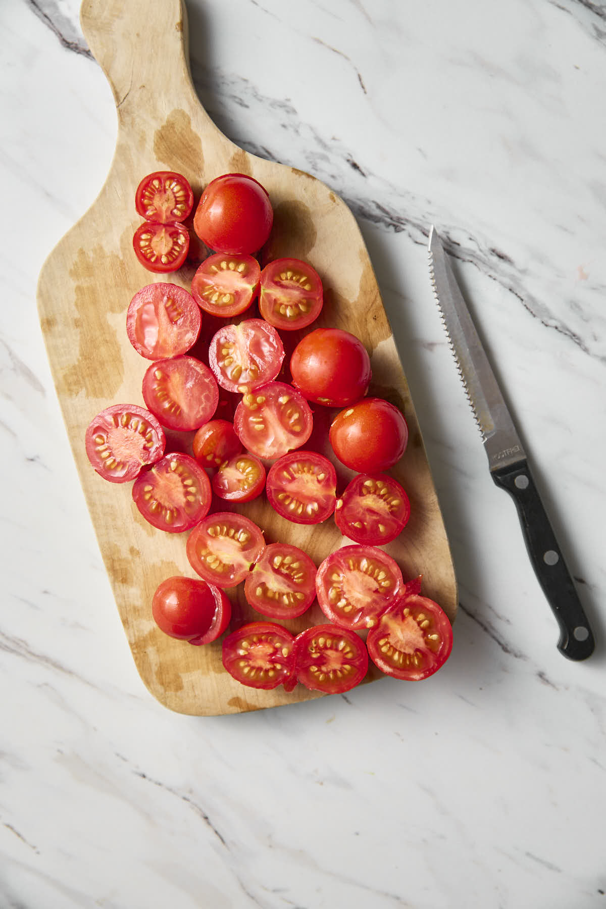Cherry tomatoes cut into halves on a wooden cutting board with a knife on the side.