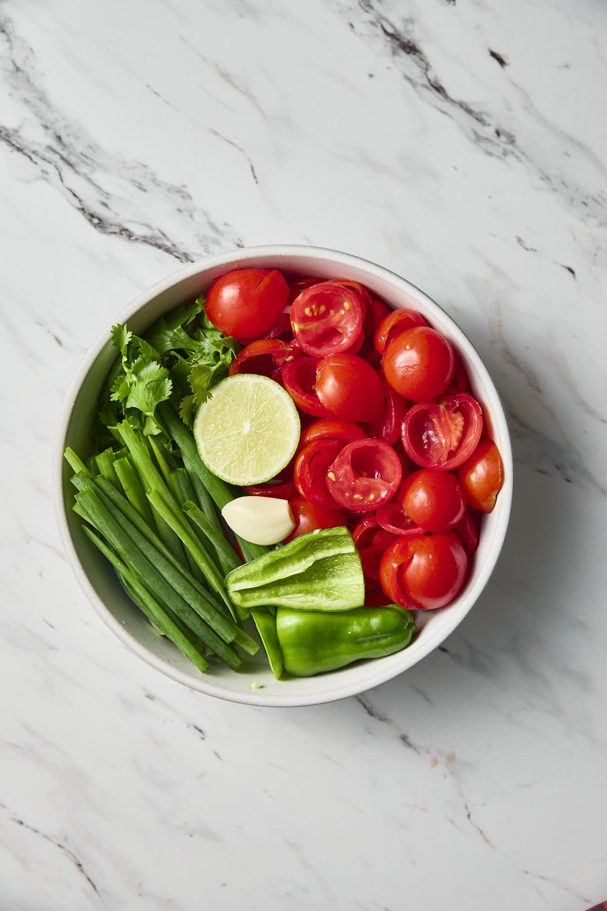 Seeded cherry tomatoes and jalapeno, garlic clove, chopped scallions, cilantro and half lime placed in a white ceramic bowl.