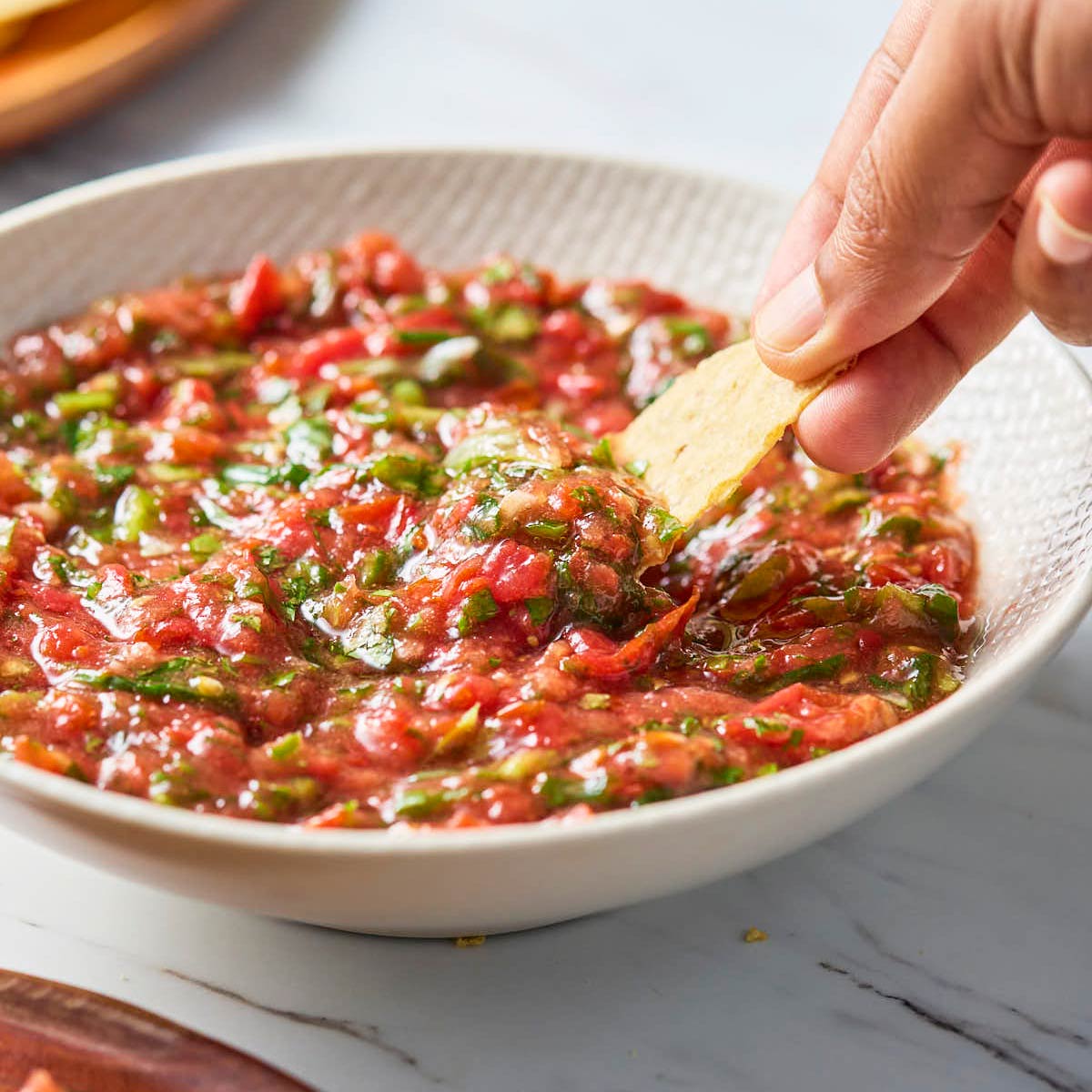 Cherry tomato salsa in a white serving bowl and someone scooping it with a tortilla chip.
