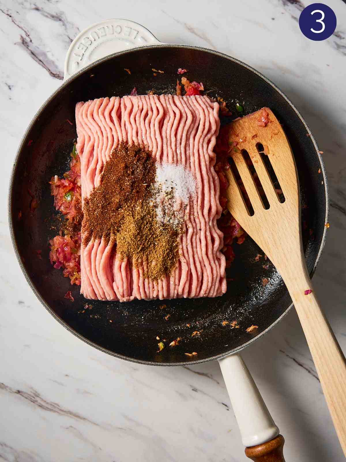 Ground turkey with spices placed on cooked onion-tomato mixture in a hot pan with wooden spatula.