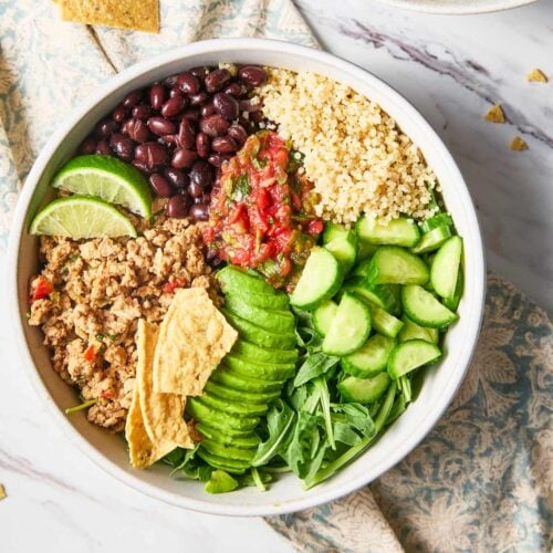 Ground turkey, beans, greens and quinoa topped with fresh cucumber, avocado and salsa in a serving bowl to serve as a burrito bowl.