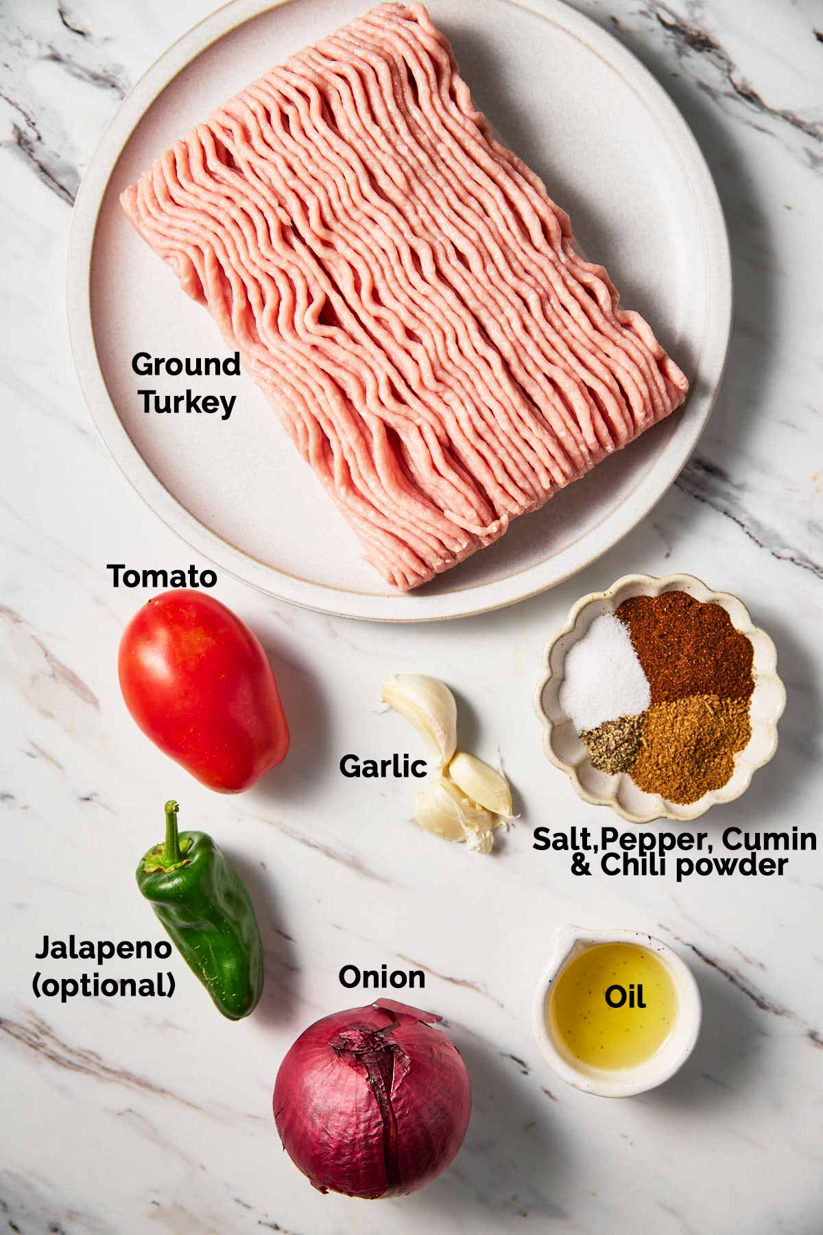 Ground turkey in a small plate, seasonings in small bowl, tomato, garlic cloves, onion, oil and jalepano placed on a flat surface for making ground turkey burrito bowl.