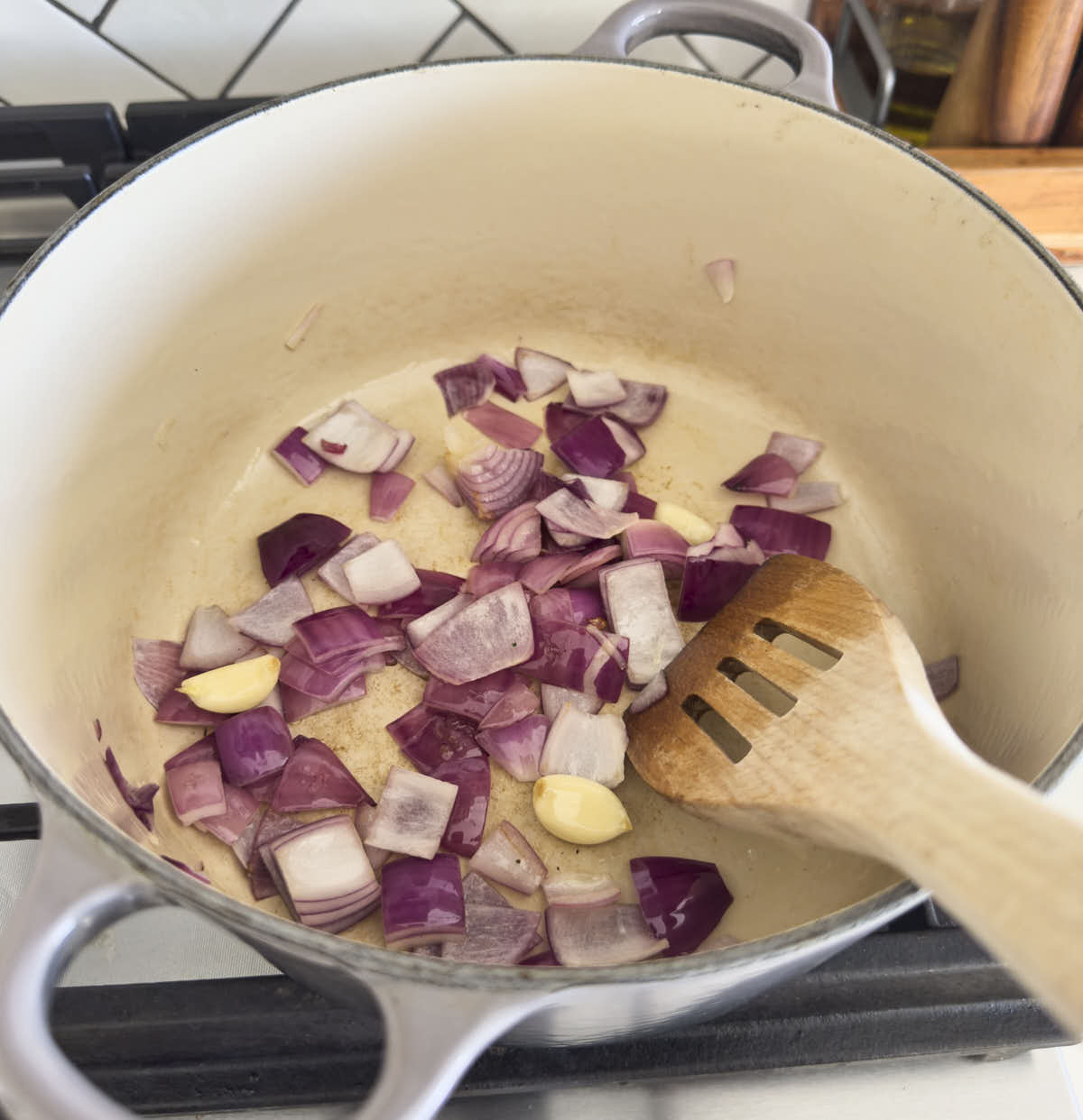 Sautéing onion and garlic cloves in a cast iron dutch oven with a wooden spatula.