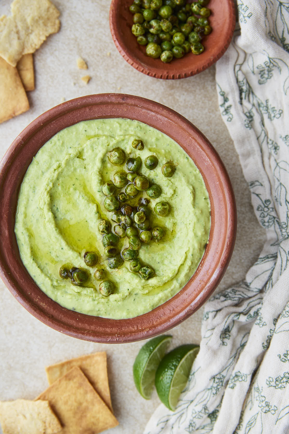 Green pea hummus in a terracotta bowl, topped with roasted green peas and a drizzle of olive oil placed on a flat surface with pita chips, lime wedges, and some more roasted peas.