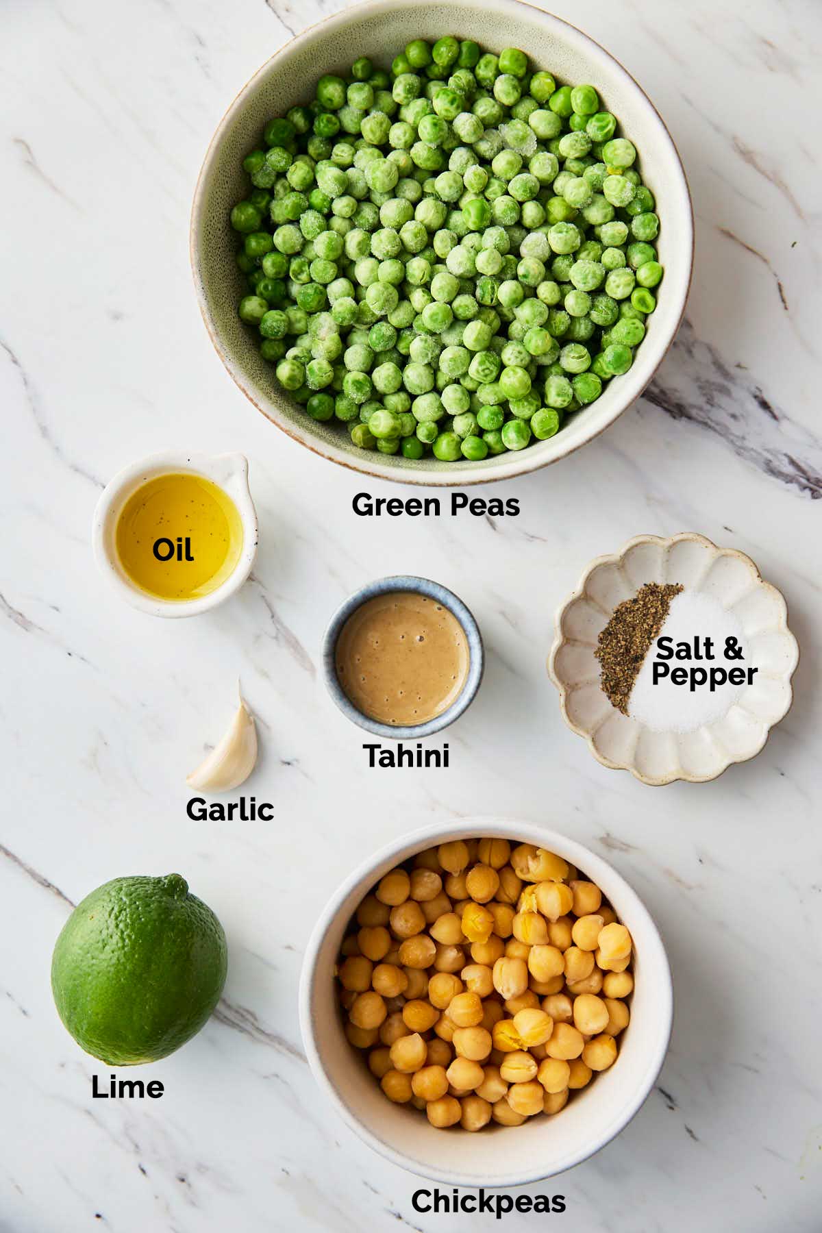 Ingredients for homemade hummus, including fresh green peas, chickpeas, tahini, lime, garlic, and olive oil, arranged on a flat surface.