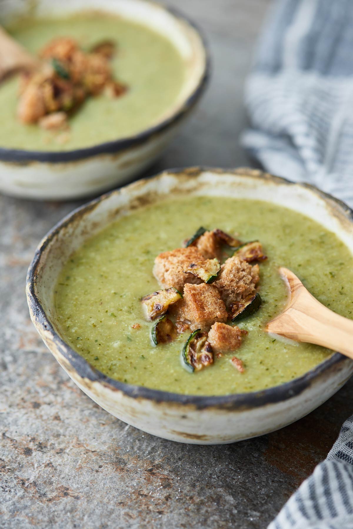 Broccoli and zucchini soup in a ceramic serving bowl with a wooden spoon. Garnished with bread croutons and roasted zucchini pieces.