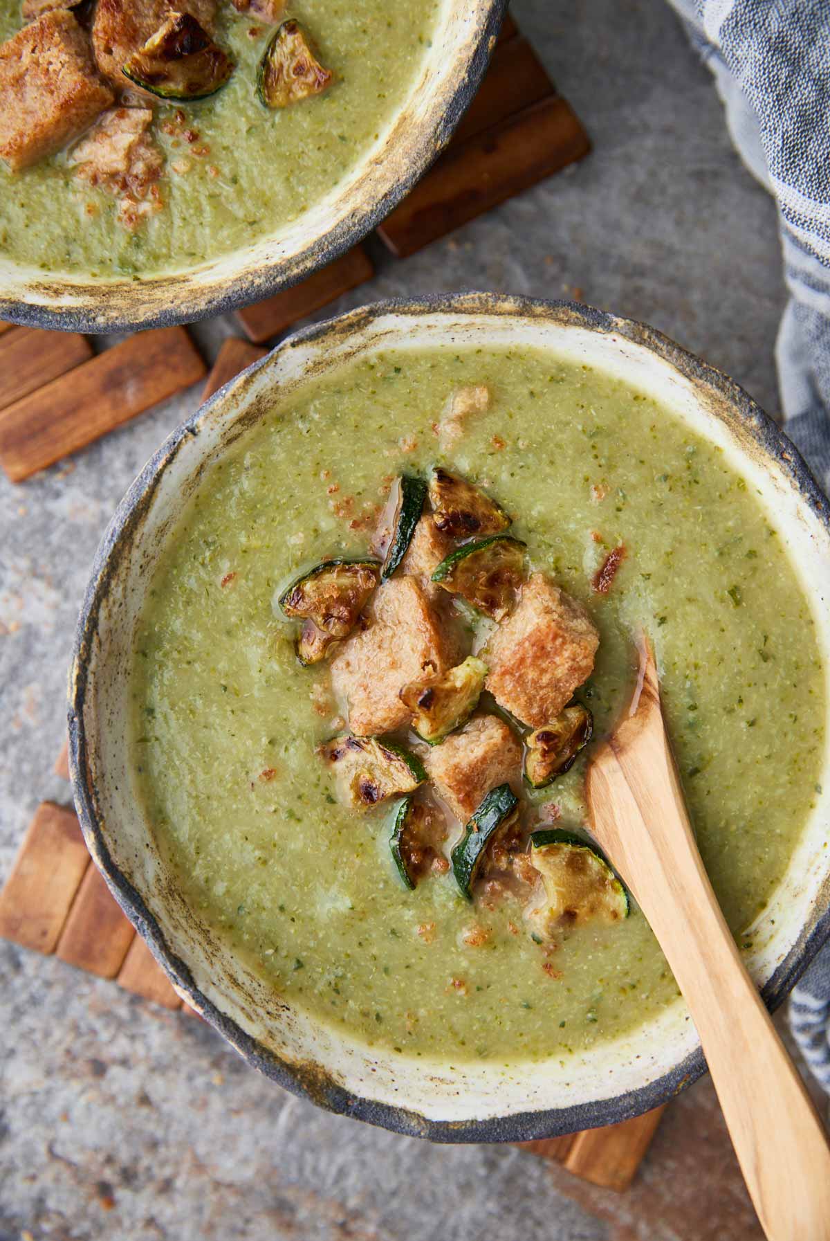 Broccoli and zucchini soup in a ceramic serving bowl with a wooden spoon. Garnished with bread croutons and roasted zucchini pieces.