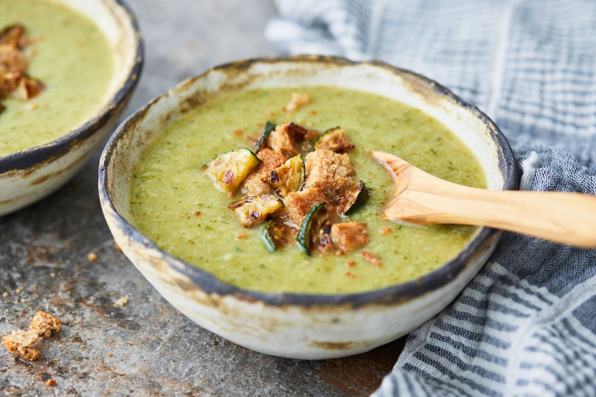 Broccoli and zucchini soup in a ceramic serving bowl with a wooden spoon. Garnished with bread croutons and roasted zucchini pieces.