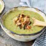 Broccoli and zucchini soup in a ceramic serving bowl with a wooden spoon. Garnished with bread croutons and roasted zucchini pieces.