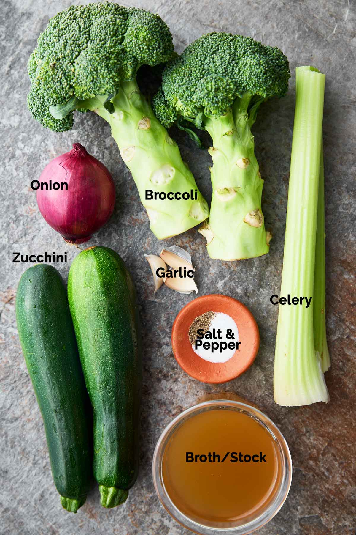 Fresh ingredients for broccoli and zucchini soup, including broccoli florets, zucchini, garlic, onion, celery, broth and seasonings on a flat surface.