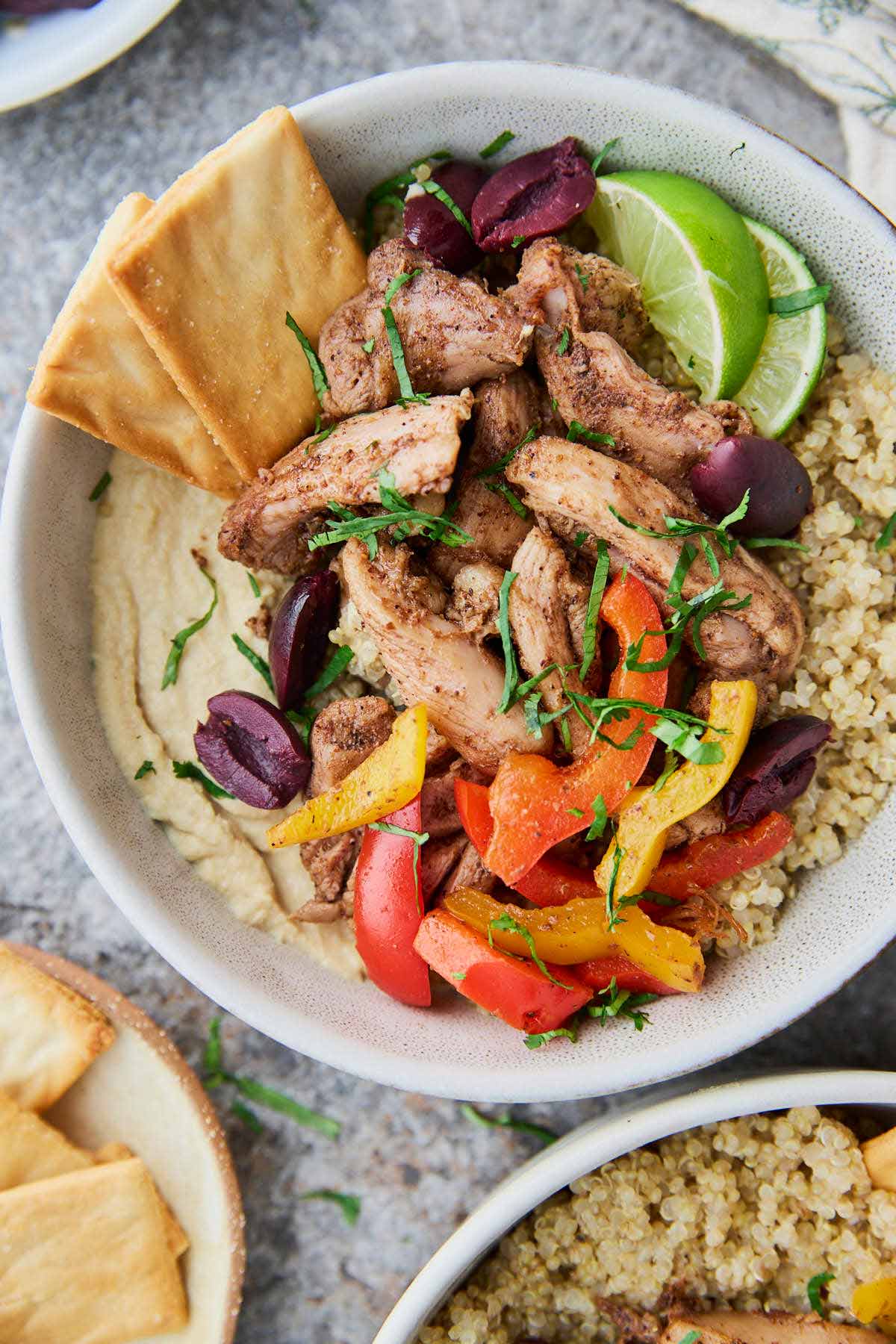 Assembled roasted shawarma chicken hummus bowl with quinoa and peppers, topped with fresh herbs, olives and lime wedges.