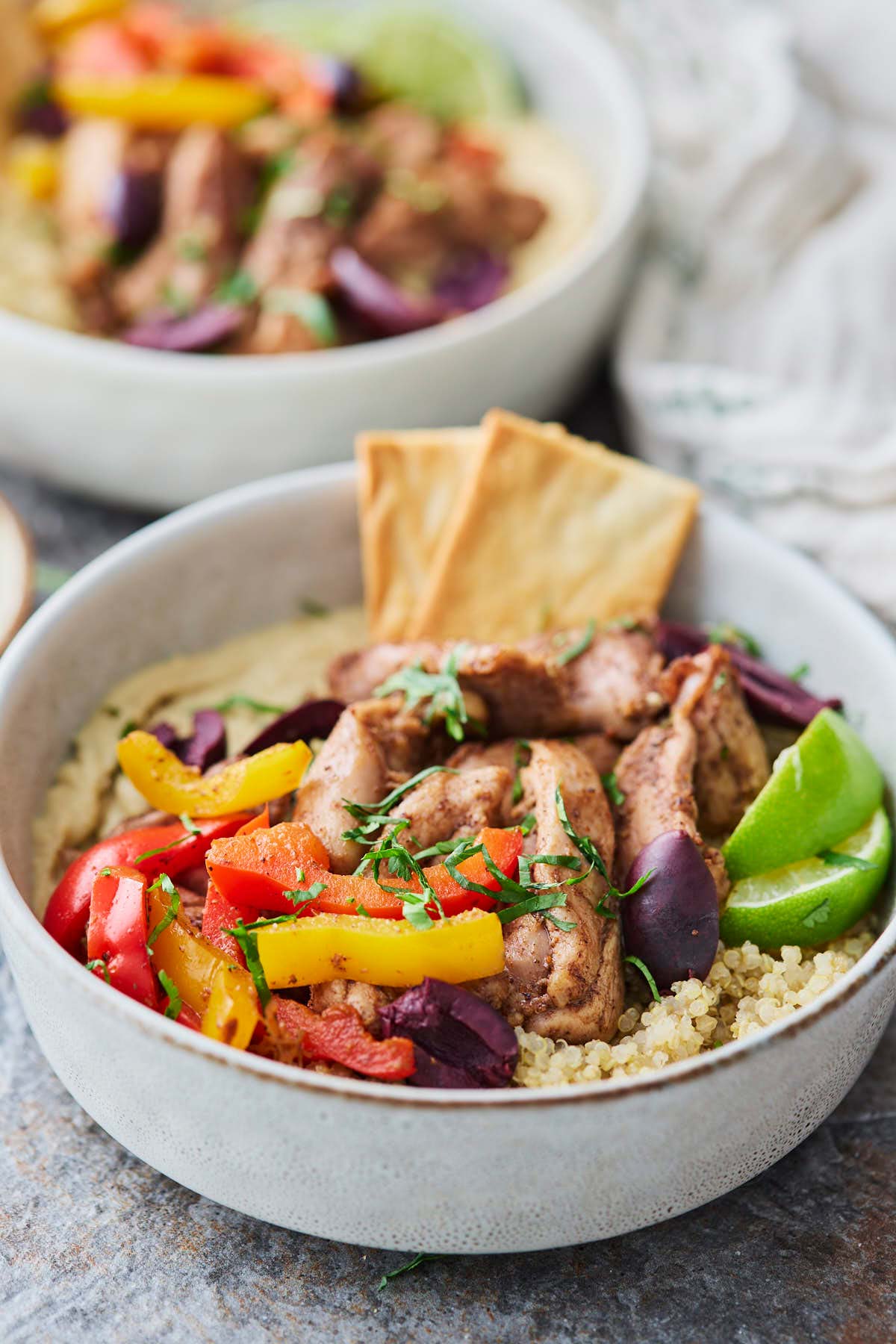 Assembled roasted shawarma chicken hummus bowl with quinoa and peppers, garnished with fresh herbs, olives and lime wedges.