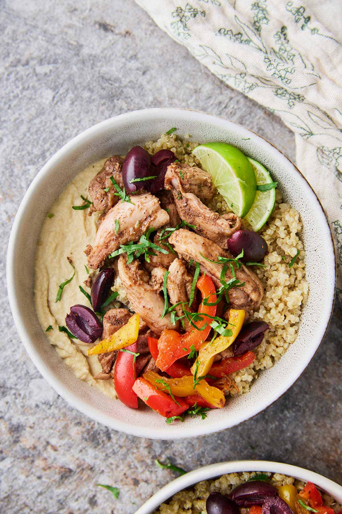 A bowl filled with shawarma chicken, hummus, quinoa, and roasted peppers, topped with fresh cilantro, olives and lime for garnish.