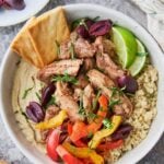 A bowl filled with shawarma chicken, creamy hummus, fluffy quinoa, and roasted peppers, topped with fresh cilantro, olives and lime for garnish.