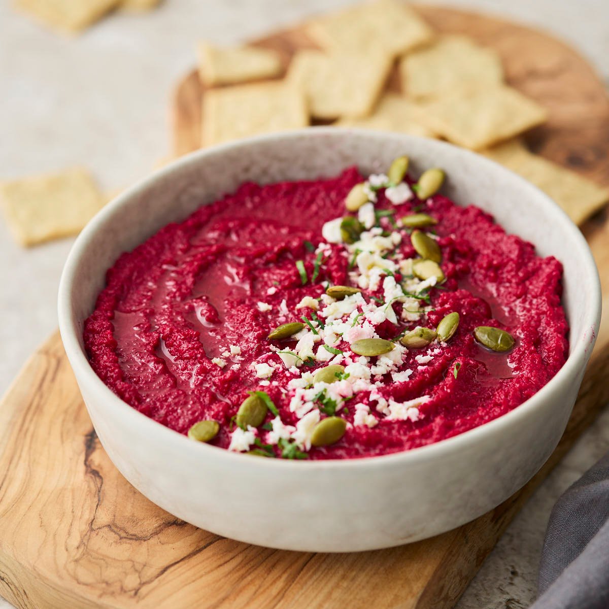 Angled view of beet and feta dip in a serving bowl on a wooden board, garnished with crumbled feta, parsley, and pumpkin seeds, alongside crackers.