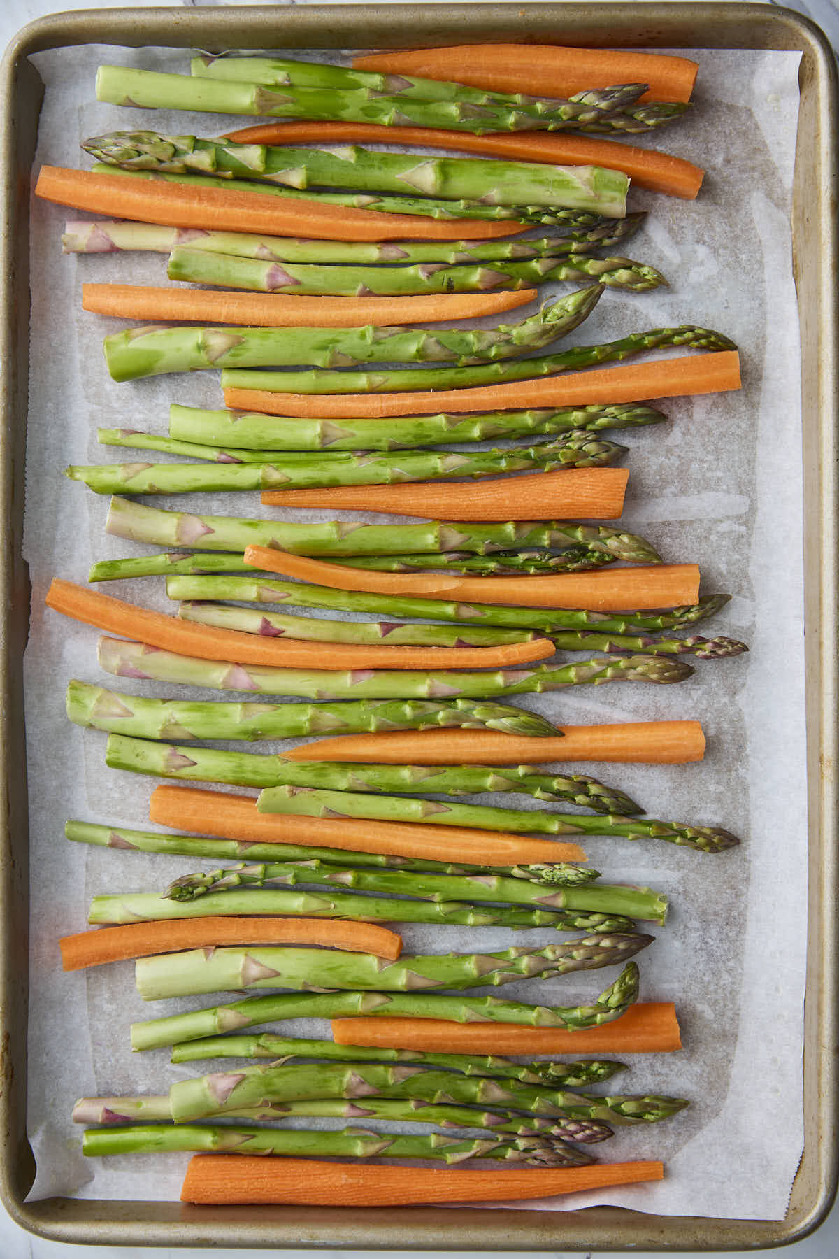 Raw asparagus spears and carrot sticks arranged in neat rows on a parchment-lined baking sheet before roasting.