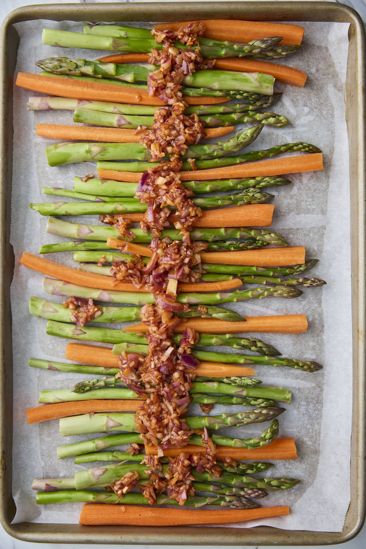 Seasoned red onion mixture spooned over raw asparagus and carrots on a baking tray, preparing them for oven-roasted asparagus and carrots.