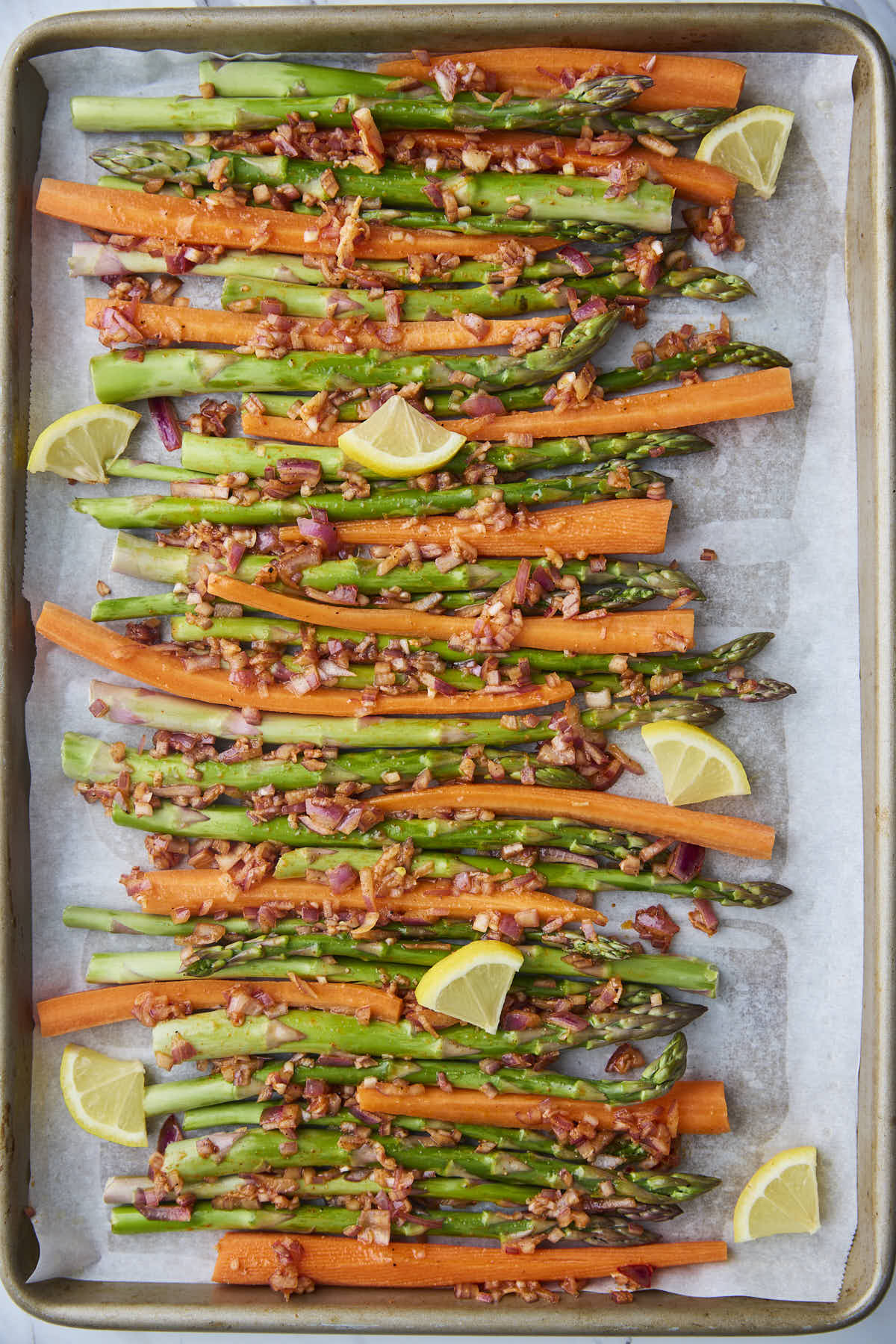 Raw asparagus and carrots evenly spread on a baking sheet with lemon wedges added for roasting, ready to go into the oven.